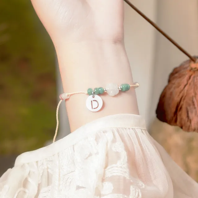 Cổ tay đeo vòng chỉ sáp màu be có hoa trà ngọc trắng, hạt ngọc xanh và charm chữ D